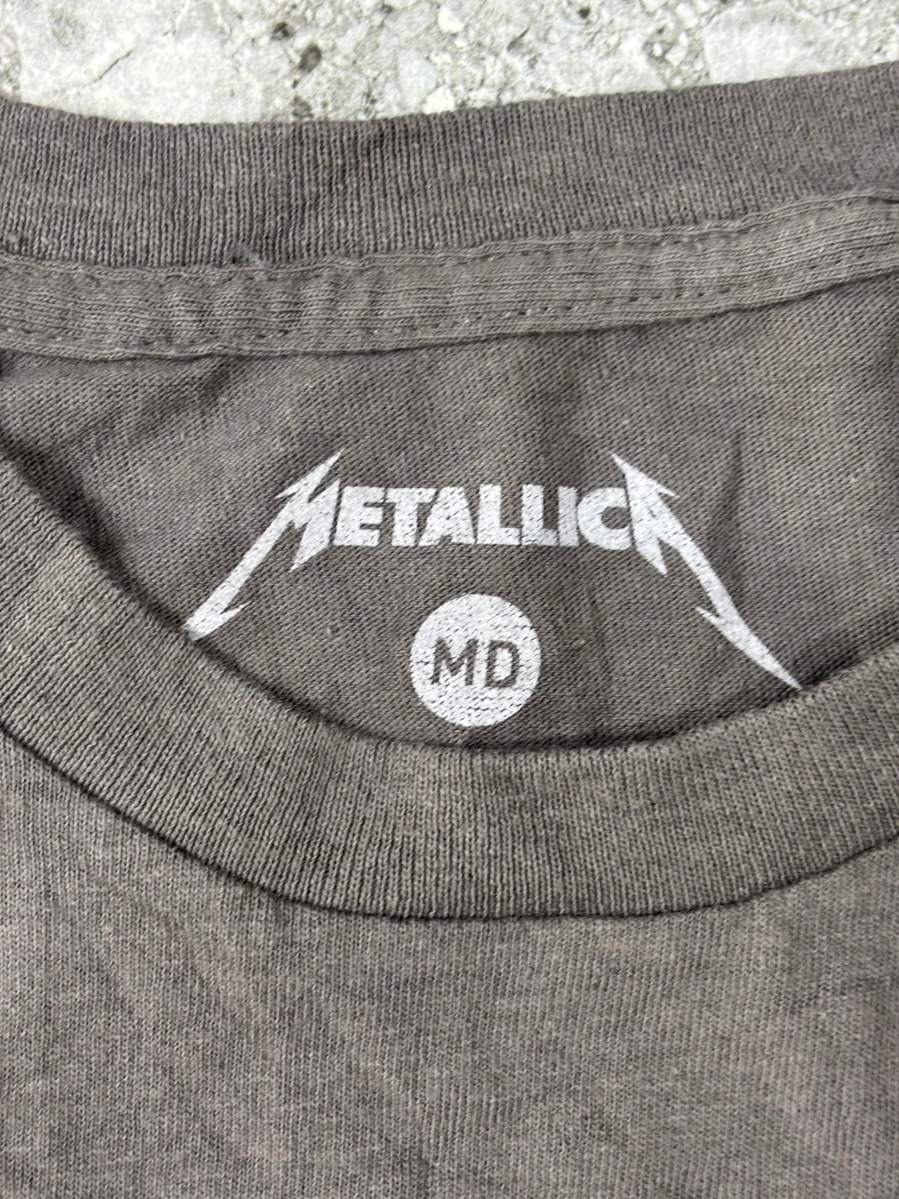 Close-up of a dark gray heather t-shirt showing the inner neck print with the white Metallica logo and 'MD' size label.